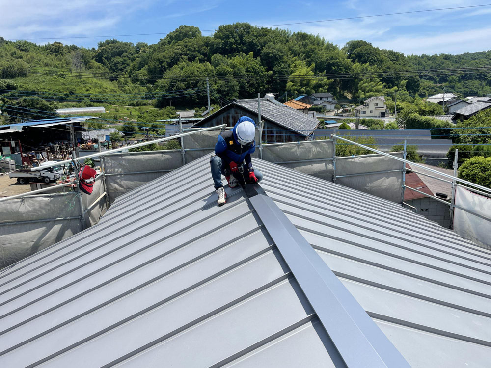 Roofer in hard hat kneels on a grey metal roof, secured by scaffolding around the building, performing installation work.