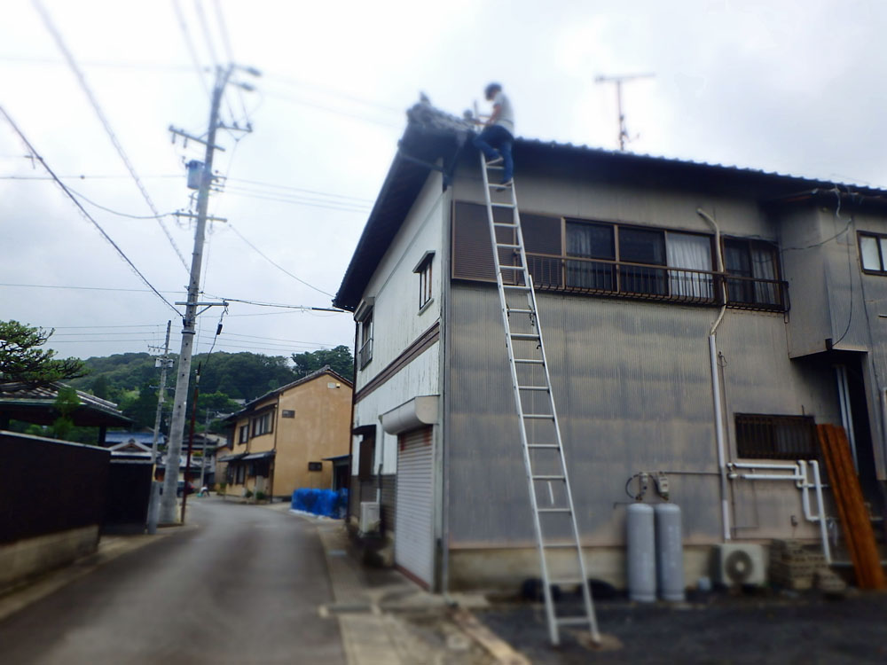 Worker on a tall ladder repairing the roof of a gray two-story building on a quiet residential street.