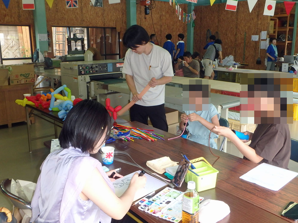 Workshop scene with people crafting at a long table; a woman draws while others braid cords and make balloon sculptures in a busy maker space.
