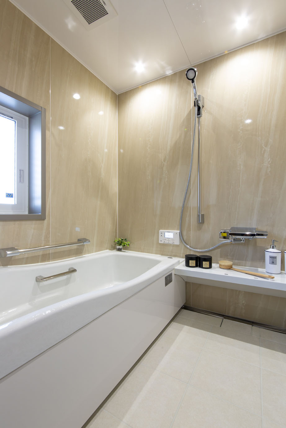 Modern beige-tiled bathroom with a white rectangular bathtub, chrome railings, and a handheld shower on the wall.
