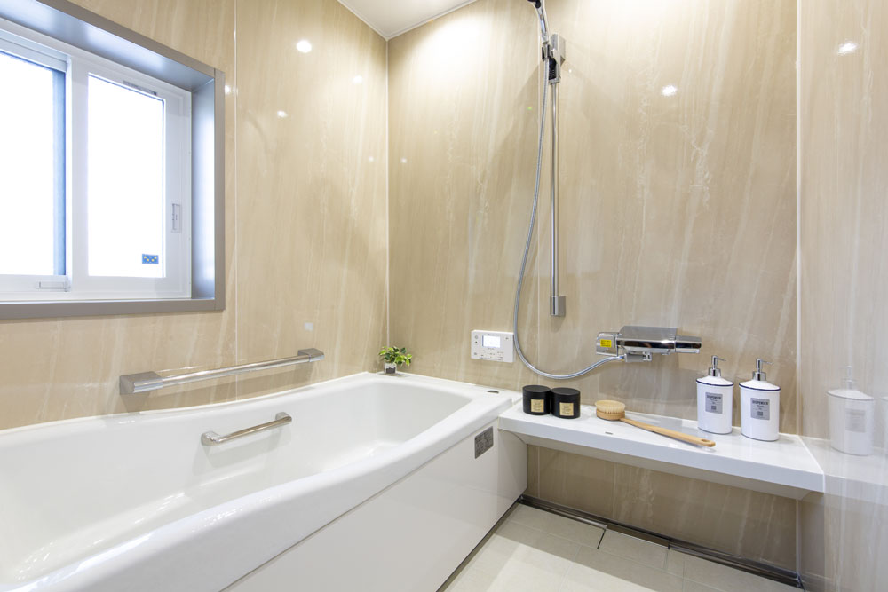 Modern bathroom with a white bathtub, beige marble walls, a window, and a bench holding toiletries and a small plant.