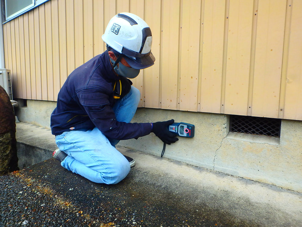 Construction worker in a safety helmet and mask crouches by a building foundation, using a handheld tester against the wall to check for moisture.